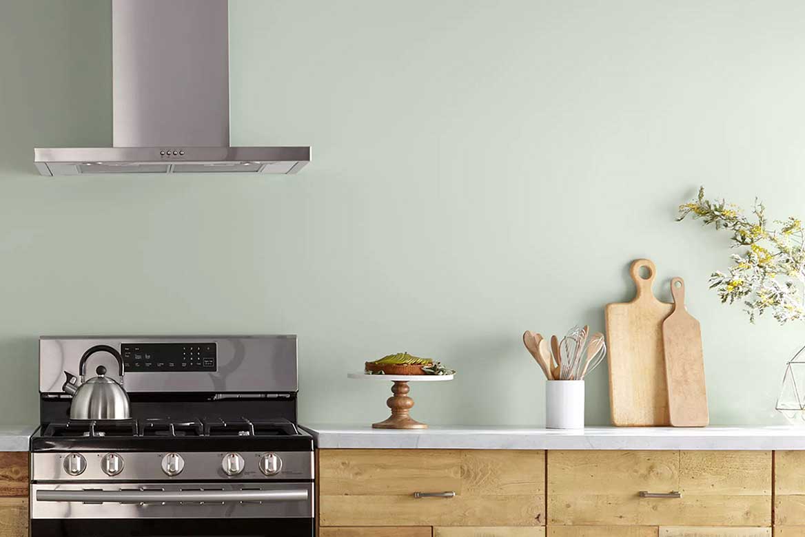 Light sage-green kitchen walls, wood cabinets, steel and black stove with kettle and cutting boards on counter.